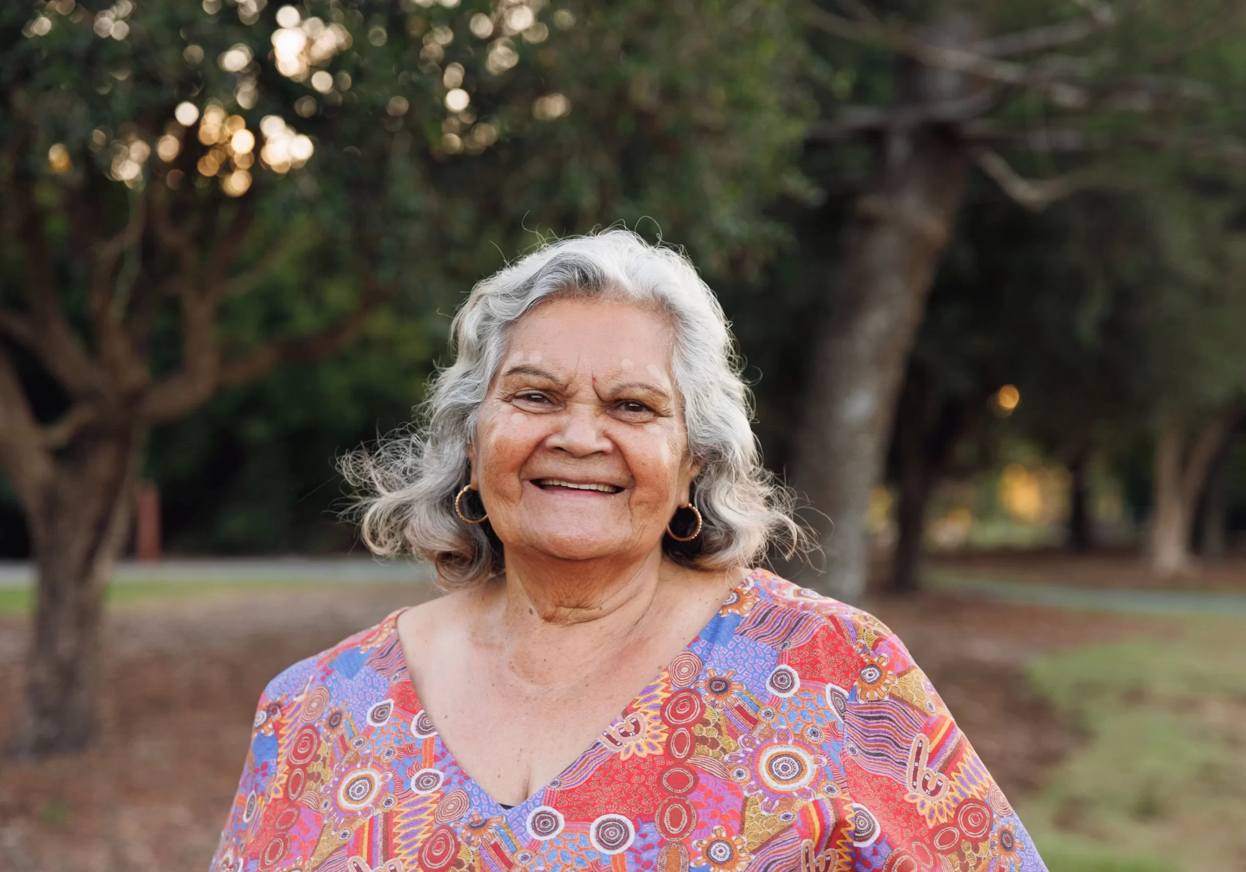 Elder indigenous women smiling at camera. Wear brightly coloured shirt.